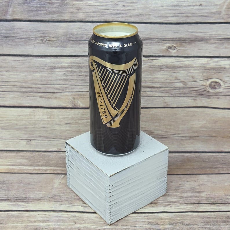 May include: A black and gold Guinness-branded candle in a can shape, with the harp logo and "ESTD 1759" printed on the front. The candle sits on a white square block against a wooden background.
