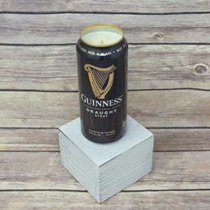 May include: A black Guinness Draught Stout can candle with a gold rim and a white wax interior. The can sits on a white square block against a light wood background. The Guinness logo is prominently displayed.