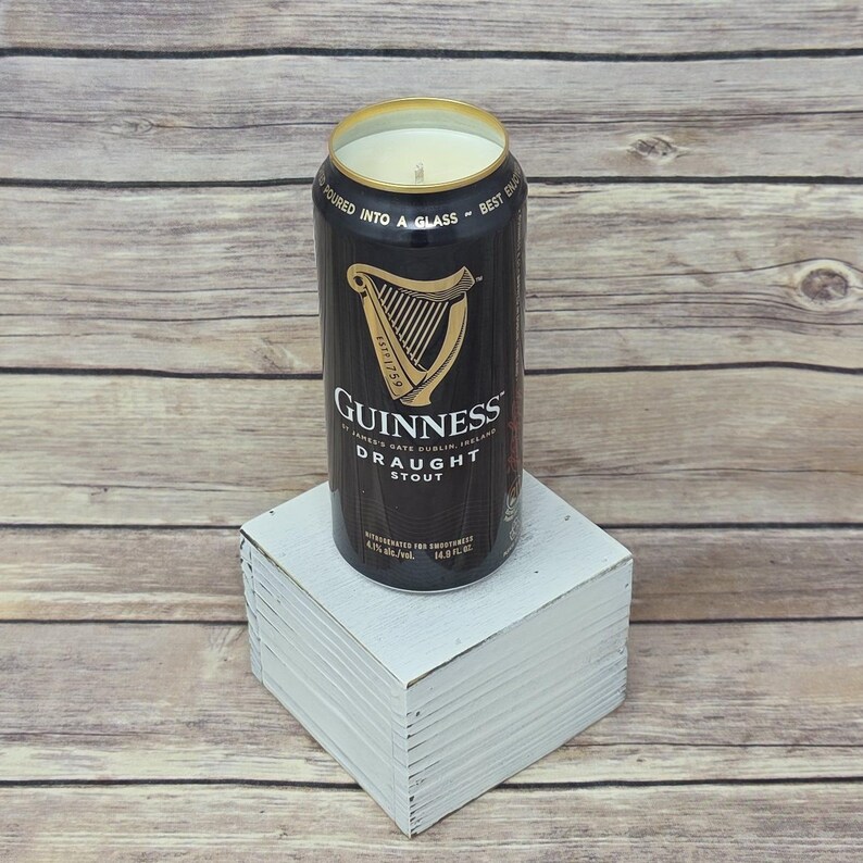 May include: A black Guinness Draught Stout can candle with a gold rim and a white wax interior. The can sits on a white square block against a light wood background. The Guinness logo is prominently displayed.