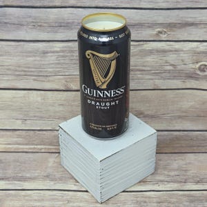 May include: A black Guinness Draught Stout can candle with a gold rim, sitting on a white block. The can features the Guinness logo and text. The candle is a unique home decor item.