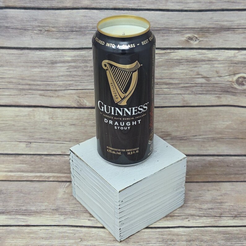 May include: A black Guinness Draught Stout can candle with a gold rim, sitting on a white block. The can features the Guinness logo and text. The candle is a unique home decor item.