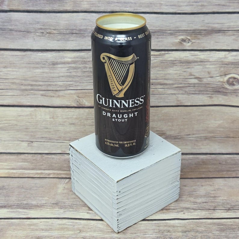 May include: A black Guinness Draught Stout can with gold lettering and a harp logo, resting on a white square block. The can reads "Guinness Draught Stout" and "4.1% alc./vol., 14.9 fl. oz."