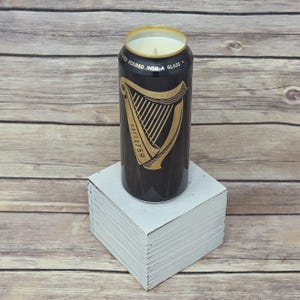 May include: A black candle in a repurposed Guinness can, featuring a gold harp design and the text "ESTD 1759." The top of the can has a gold rim and the text "POURED INTO A GLASS."