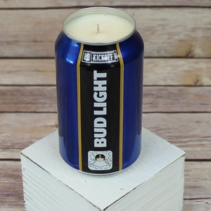 May include: A blue and gold Bud Light can candle with the text "Kickoff" and a football graphic. The candle is sitting on a white wooden surface.