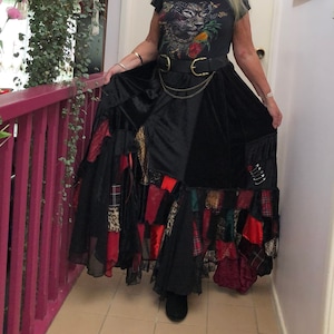 May include: A black maxi skirt with patchwork details, featuring red, plaid, and animal print fabrics. The skirt is paired with a black t-shirt with a leopard graphic and a double-buckle belt. The outfit is completed with a gold chain and black boots.