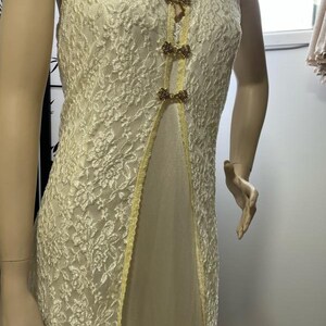 Puede incluir: Vestido sin mangas de encaje marfil con cuello en V. El vestido presenta una capa transparente con estampados florales y está acentuado con cierres dorados en la parte delantera. Un ribete amarillo bordea la abertura.