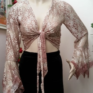 May include: A sheer, long-sleeved, tie-front top in a blush pink hue, adorned with intricate floral lace. The top features flared sleeves and is paired with a black skirt. The mannequin is displayed in a well-lit setting.