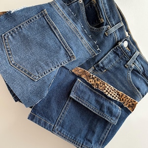 May include: Close-up of denim shorts with a patchwork design. The shorts are a medium wash blue with a leopard print accent. The shorts have a raw hem and a cargo pocket.