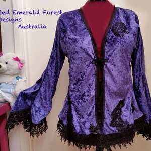 Op de afbeelding: Een paarse fluwelen jas met lange, uitlopende mouwen en zwarte kanten afwerking. De jas heeft een V-hals, zwarte knoopsluitingen en decoratieve zwarte kanten applicaties. De tekst "Enchanted Emerald Forest Designs Australia" is zichtbaar.