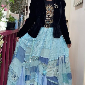 May include: A woman wearing a black velvet jacket over a graphic tee and a long, patchwork skirt in shades of blue. The skirt features lace and floral patterns. The website address www.enchanted-emerald-forest-designs.com is visible.