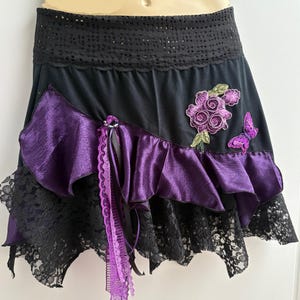 May include: Black mini skirt with a black lace hem and a purple satin ruffle. The skirt features a purple floral applique and a butterfly applique. A purple lace and ribbon detail hangs from the waist.