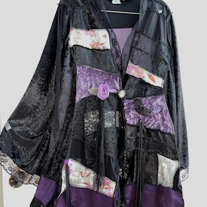 Könnte beinhalten: Lange Patchwork-Jacke aus schwarzem Samt, Spitze und lila Satin. Die Jacke hat florale Akzente, dekorative Knöpfe und Spitzensaum. Sie hängt an einem schwarzen Kleiderbügel vor weißem Hintergrund.
