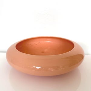 May include: A peach-colored, rounded bowl with a glossy finish. The bowl has a wide, shallow design and sits on a white surface. The interior of the bowl is the same color as the exterior.