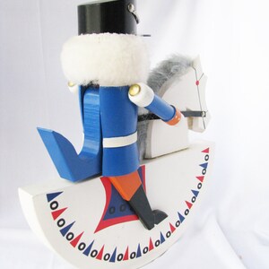 May include: A wooden toy nutcracker soldier in a blue uniform and black hat, riding a white rocking horse. The rocking horse has a gray mane and tail. The base is white with red and blue decorative accents.