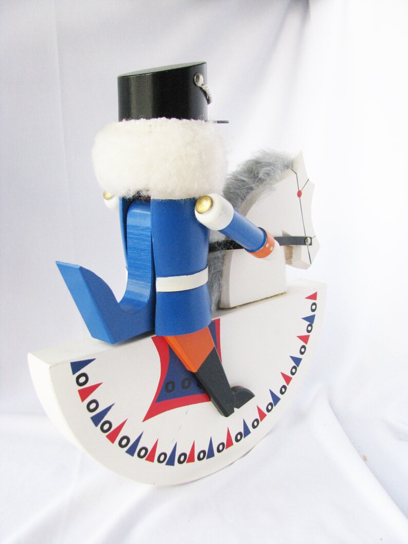 May include: A wooden toy nutcracker soldier in a blue uniform and black hat, riding a white rocking horse. The rocking horse has a gray mane and tail. The base is white with red and blue decorative accents.