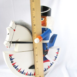 May include: A wooden nutcracker soldier riding a white rocking horse. The nutcracker is wearing a blue uniform and a black hat. The rocking horse is decorated with red and blue accents. The nutcracker is approximately 11 inches tall.