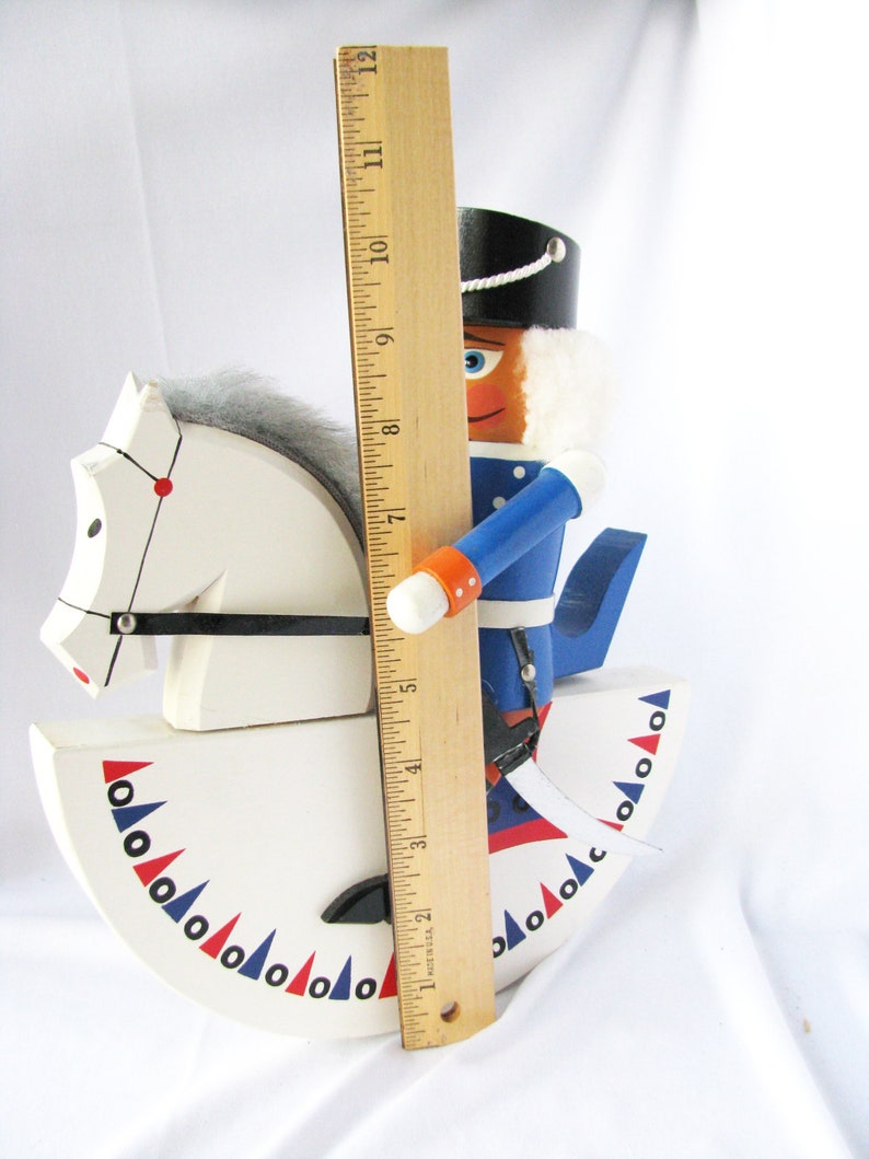 May include: A wooden nutcracker soldier riding a white rocking horse. The nutcracker is wearing a blue uniform and a black hat. The rocking horse is decorated with red and blue accents. The nutcracker is approximately 11 inches tall.