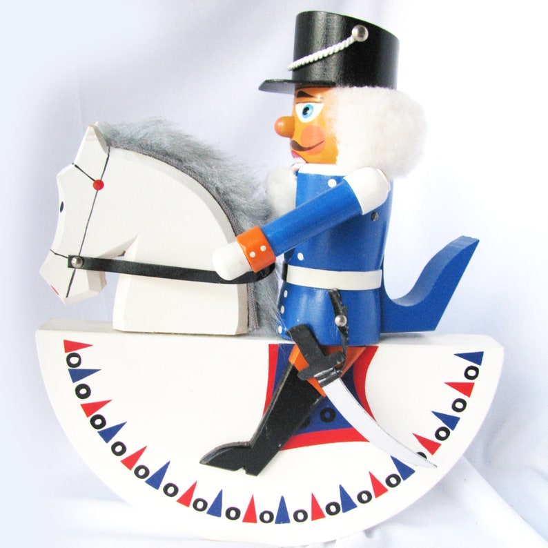 May include: A wooden toy soldier riding a white horse on a rocking base. The soldier wears a blue uniform with white trim, a black hat, and a white fur collar. The rocking base is white with red and blue triangles.