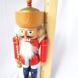 May include: A wooden nutcracker figurine with a red jacket, blue boots, and a white beard. The figurine has a wooden bowl on its head and is holding a golden staff. The figurine is approximately 23 cm tall.