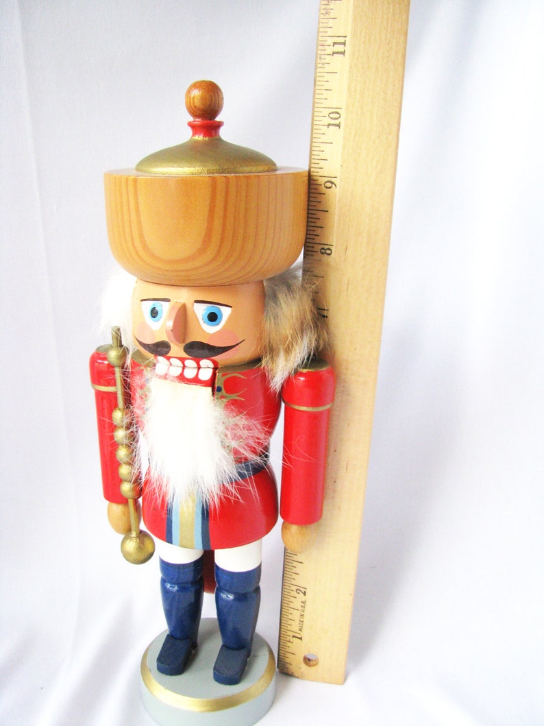 May include: A wooden nutcracker figurine with a red jacket, blue boots, and a white beard. The figurine has a wooden bowl on its head and is holding a golden staff. The figurine is approximately 23 cm tall.
