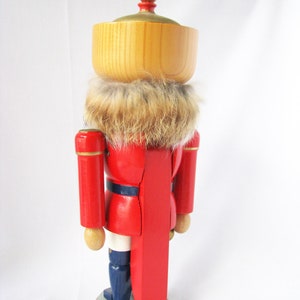 May include: A wooden nutcracker figurine in a red uniform with a faux fur collar. The figurine has a light brown wooden head and a gold-coloured top. The base is light grey with a gold trim, and the boots are navy blue.