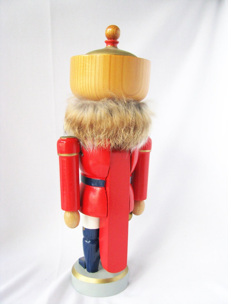 May include: A wooden nutcracker figurine in a red uniform with a faux fur collar. The figurine has a light brown wooden head and a gold-coloured top. The base is light grey with a gold trim, and the boots are navy blue.