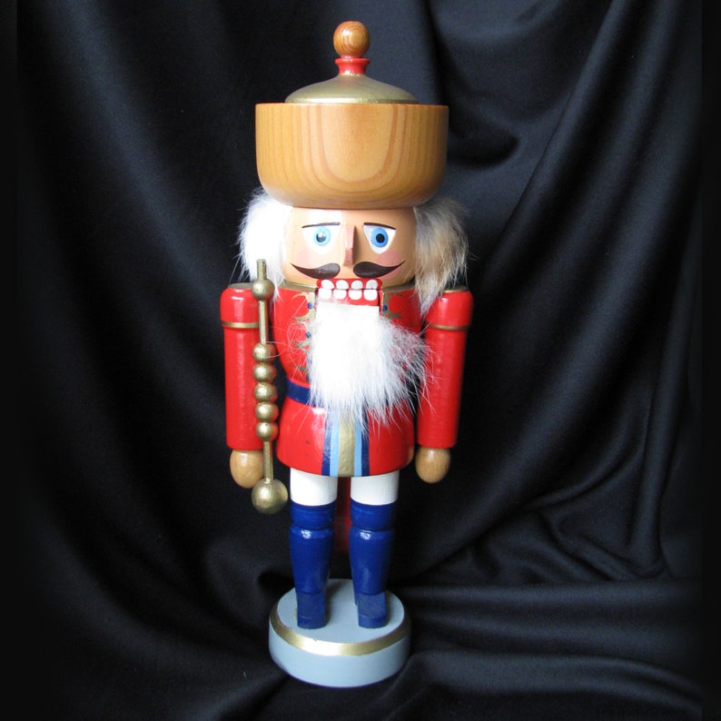 May include: A wooden nutcracker figurine in a red coat with blue trousers and a white beard. The nutcracker has a wooden bowl on its head and holds a golden staff. The figurine stands on a gray and gold base.