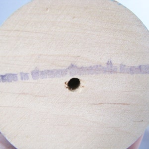 May include: Close-up of the base of a light-coloured wooden spool. A small hole is drilled in the centre. Faint purple text is stamped on the wood, reading "Made in Germany". The wood grain is visible.