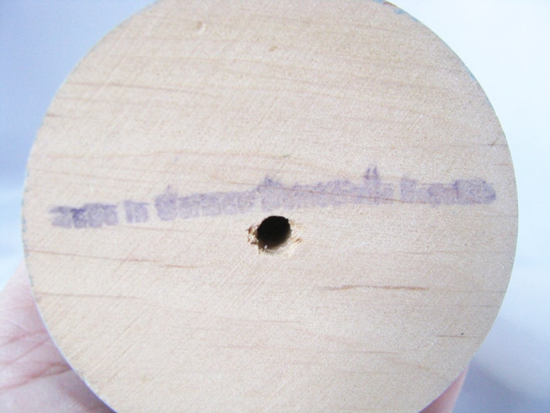 May include: Close-up of the base of a light-coloured wooden spool. A small hole is drilled in the centre. Faint purple text is stamped on the wood, reading "Made in Germany". The wood grain is visible.