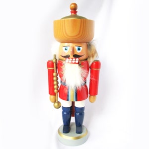 May include: A wooden nutcracker figurine. It features a red coat, blue trousers, and a large wooden hat. The figurine has a white beard, a mustache, and holds a golden staff. The base is light grey with a gold trim.