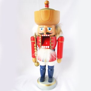 May include: A wooden nutcracker figurine. It features a red jacket, blue trousers, and a large wooden hat. The figurine has a white beard and mustache, and holds a golden staff. A classic Christmas decoration.