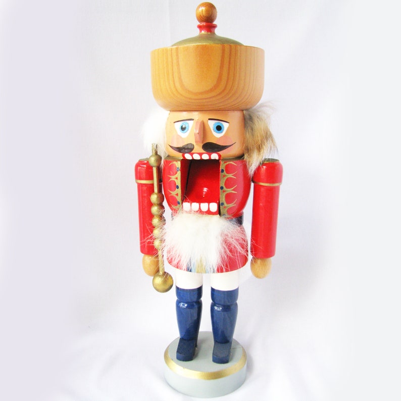 May include: A wooden nutcracker figurine. It features a red jacket, blue trousers, and a large wooden hat. The figurine has a white beard and mustache, and holds a golden staff. A classic Christmas decoration.