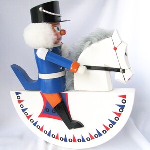 May include: A wooden toy soldier riding a white horse-shaped rocker. The soldier wears a blue uniform, black hat, and has a white beard. The rocker base is white with red, blue, and black decorative triangles.