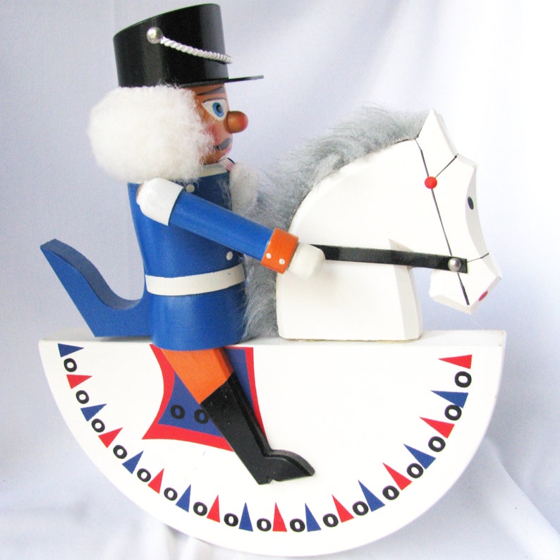 May include: A wooden toy soldier riding a white horse-shaped rocker. The soldier wears a blue uniform, black hat, and has a white beard. The rocker base is white with red, blue, and black decorative triangles.