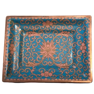 May include: Rectangular trinket dish with a turquoise background and pink floral design. The dish has a copper-colored border and features an intricate pattern of swirls and flowers. A decorative piece for holding small items.