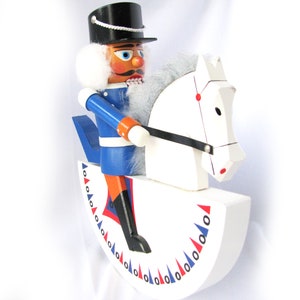 May include: A wooden nutcracker soldier riding a white rocking horse. The soldier wears a blue uniform with orange accents, a black hat, and a white wig. The rocking horse has a black bridle and red and blue decorative elements.