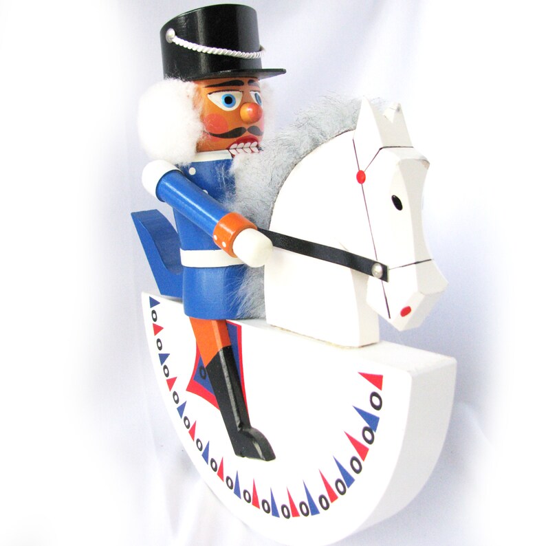 May include: A wooden nutcracker soldier riding a white rocking horse. The soldier wears a blue uniform with orange accents, a black hat, and a white wig. The rocking horse has a black bridle and red and blue decorative elements.