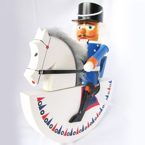 May include: A wooden nutcracker soldier riding a white rocking horse. The soldier wears a blue uniform, black boots, and a black top hat. The rocking horse has a white body with red, blue, and black accents.
