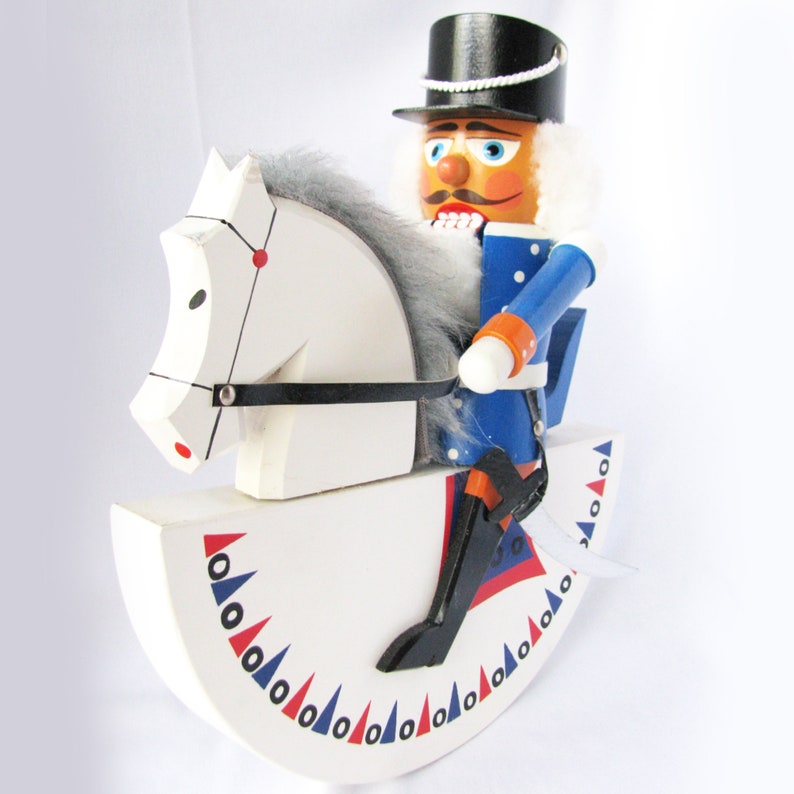 May include: A wooden nutcracker soldier riding a white rocking horse. The soldier wears a blue uniform, black boots, and a black top hat. The rocking horse has a white body with red, blue, and black accents.