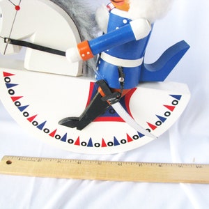 May include: A wooden rocking horse nutcracker with a white body and a blue-coated rider. The horse has a white head and a gray mane. The rider wears a blue coat with orange accents and a white fur collar. The base is decorated with red, white, and blue triangles.