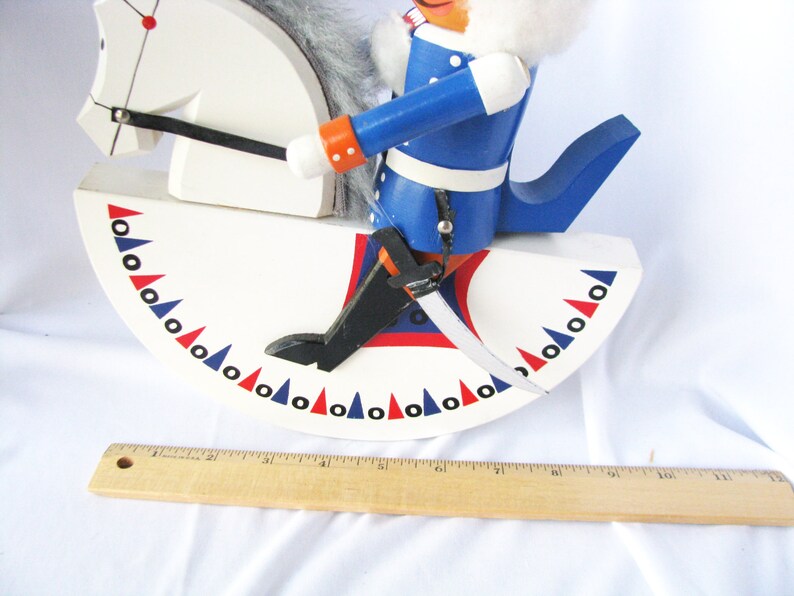 May include: A wooden rocking horse nutcracker with a white body and a blue-coated rider. The horse has a white head and a gray mane. The rider wears a blue coat with orange accents and a white fur collar. The base is decorated with red, white, and blue triangles.