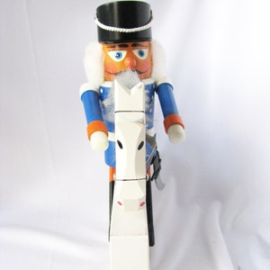 May include: A wooden nutcracker soldier with a black hat, blue jacket, and white fur trim. The nutcracker is holding a white horse-shaped object. The soldier has an orange face, blue eyes, and a white mustache.