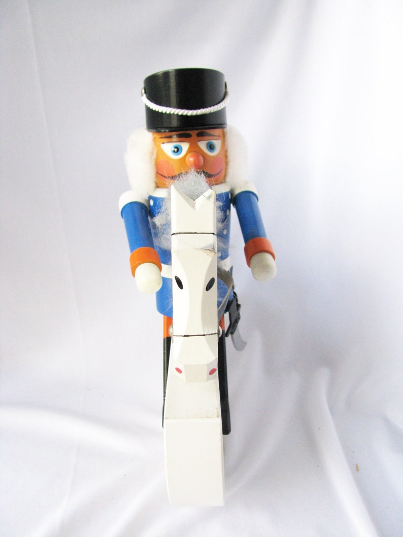 May include: A wooden nutcracker soldier with a black hat, blue jacket, and white fur trim. The nutcracker is holding a white horse-shaped object. The soldier has an orange face, blue eyes, and a white mustache.