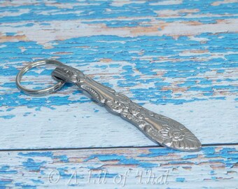 May include: A silver spoon keychain with a decorative floral design. The keychain is on a blue and white wooden background.