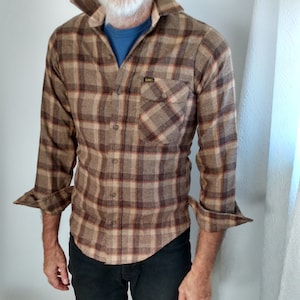 Puede incluir: Una camisa de franela a cuadros de color marrón y beige de manga larga con un solo bolsillo en el pecho. La camisa tiene una etiqueta que dice "Lee" en el bolsillo.