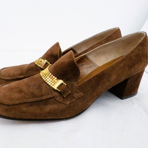 May include: Brown suede heeled loafers with a gold chain detail across the vamp.
