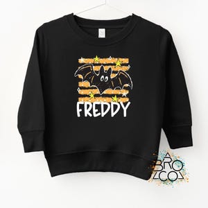 May include: A black long-sleeve sweatshirt featuring a graphic of a black bat centered over horizontal orange stripes and yellow stars. The name FREDDY is printed in white capital letters below the bat. The design has a hand-drawn aesthetic.