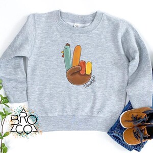 Może przedstawiać: Jasnoszara bluza z kolorowym gestem dłoni indyka i napisem "Thankful". Projekt zawiera brązowy, pomarańczowy, ż&oacute;łty, zielony i niebieski. Bluza jest zestawiona z niebieskimi dżinsami i brązowymi butami.