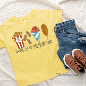 May include: Yellow t-shirt with a graphic of popcorn, peanuts, a snow cone, and a corn dog. The text on the shirt reads "I'm here for the concession stand."