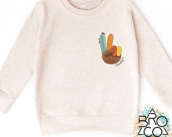 May include: A light beige sweatshirt with long sleeves. The front features a colorful graphic of a hand in a peace sign, designed to look like a turkey. The word "Thankful" is written in script. The sweatshirt is a children's apparel item.